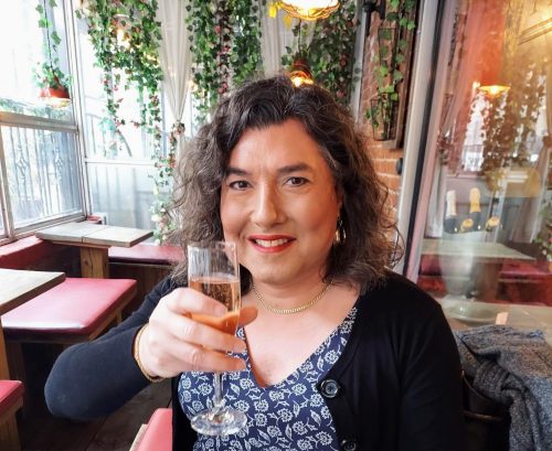 The author, wearing a dress with a white on blue floral pattern, a black sweater and gold-plated earrings and necklace, lifts a glass of prosecco to the camera