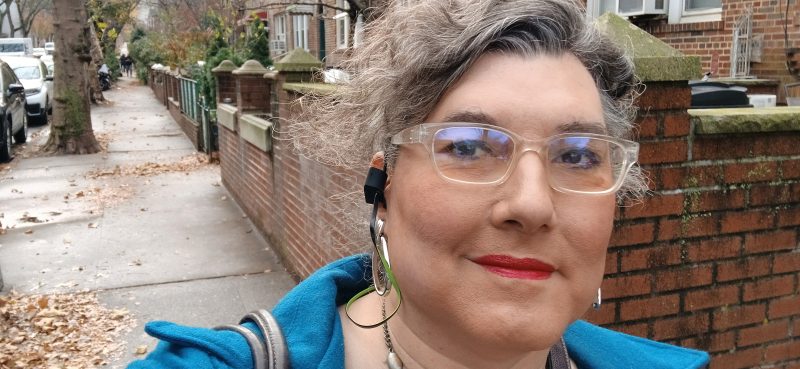 Selfie of a woman walking down the street, wearing makeup, jewelry, glasses, earbuds and a blue wool coat.
